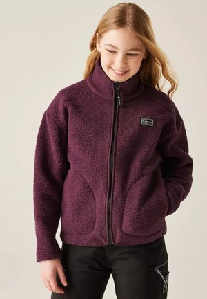 Niña sonriente con pelo largo que lleva una chaqueta de forro polar morada con cremallera y pantalones negros, con las manos en los bolsillos, sobre un fondo liso y claro.