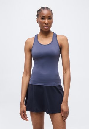 Žena s vrkočmi, oblečená v priliehavom modrom tielku bez rukávov a tmavomodrej športovej sukni, stojaca pred jednoduchým svetlým pozadím.