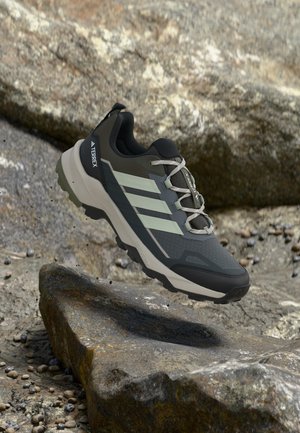 Zapatilla de senderismo Adidas Terrex negra y gris con rayas blancas, flotando sobre terreno rocoso con pequeños guijarros dispersos alrededor.