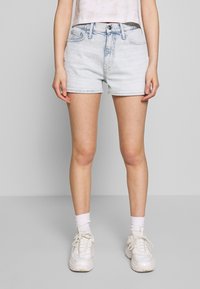 Lichtblauwe denim shorts met een hoge taille, voorzien van zijzakken en een ruwe zoom. Gecombineerd met witte sneakers en enkelsokken.