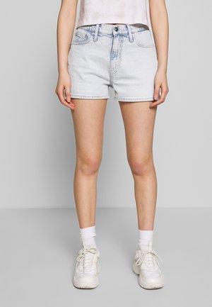 Shorts en denim bleu clair avec une taille haute, présentant des poches avant et un ourlet brut. Associés à des baskets blanches et des chaussettes hautes.