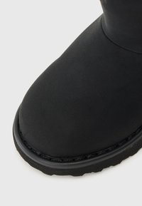 Fekete bokacsizma sima velúrból, lekerekített orral és texturált gumitalppal. A felső élen végig varrott varrás található.