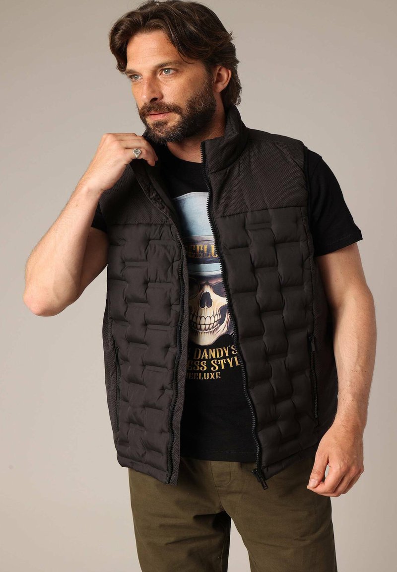 Veste noire rembourrée avec fermeture éclair à l'avant, dotée d'un design matelassé. Portée sur un t-shirt noir graphique et assortie avec un pantalon olive.