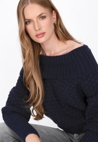 Maglione in maglia blu navy con una trama textured e lavorazione a coste sui polsini. Presenta un scollo a spalla scoperta. Il modello è seduto e indossa pantaloni grigi.