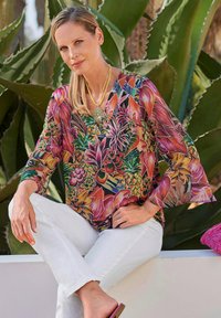 Losse blouse met levendige bloemen- en fruitpatronen in roze, groen en geel; gecombineerd met witte broek en een gouden ketting.