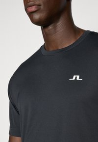Sort atletisk t-shirt lavet af glat stof, med kortærmet design og et hvidt logo på brystet. Enkel og strømlinet.