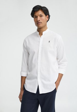 Uomo con camicia bianca con collo alla mandarina, maniche rimboccate e pantaloni scuri, in piedi con una mano in tasca.
