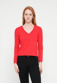 Suéter rojo de cuello en V con mangas largas, puños acanalados y dobladillo recortado. Hecho de un material de punto suave, combinado con pantalones negros.