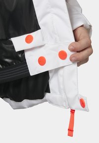 Bílá sekce bundy s černým interiérem, obsahující oranžové patentky a červené poutko. Texturovaná látka je viditelná ve detailech.
