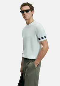 Man in zwarte zonnebril, witte korte mouwen shirt met zwarte strepen op de mouwen, en olijfgroene broek, met een zwarte tas in de hand.