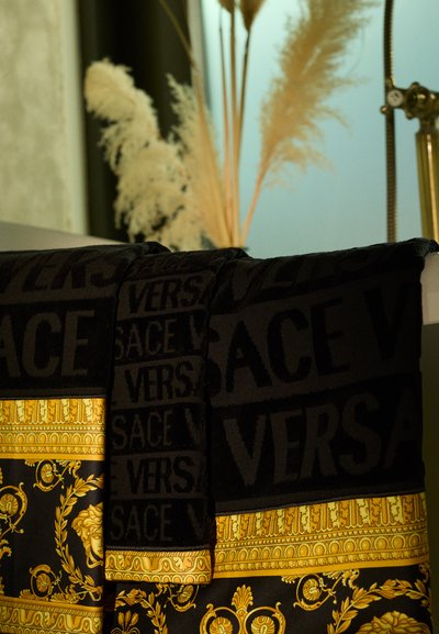 Juoda tekstilė su iškiliu užrašu "VERSACE" ir auksiniais raštais, sudėtingais dizainais kontrastingame fone.