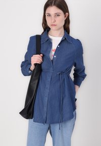 Camisa de ganga em azul escuro com uma cintura ajustável, apresentando colarinho, mangas longas e fecho à frente com botões. Combinada com jeans azul claros e uma bolsa preta.