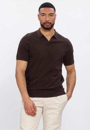Mann mit kurzen Haaren und Bart, der ein dunkelbraunes kurzärmeliges Poloshirt und hellbeige Hosen trägt, steht vor einem einfarbigen weißen Hintergrund.