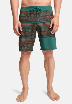 Man die groene en bruine zwemshorts met tribale patronen tot op de knie draagt en een voorstrik heeft, staand op blote voeten tegen een effen achtergrond.