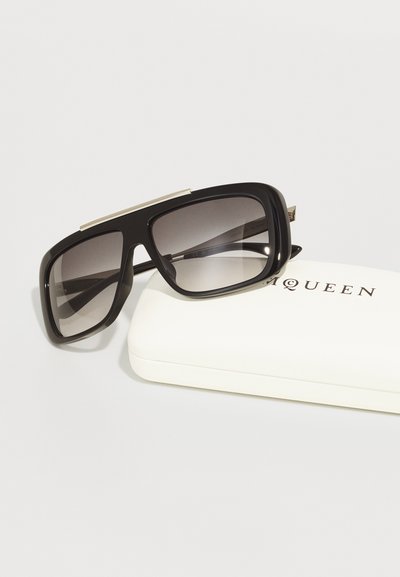 Black oversized sunglasses with gradient lenses, rectangular shape, and metal accent on the top. Accompanied by a white protective case.
