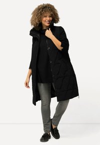 Mustat tikatulle pitkä takki, jossa on korkea kaulus, vetoketjukiinnitys ja napin yksityiskohtia, yhdistettynä mustaan neuleeseen ja harmaisiin farkkuihin.