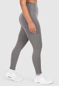Leggings grigi alti in vita realizzati in tessuto elasticizzato, caratterizzati da una superficie liscia e dettagli di cucitura sottili, abbinati a scarpe da ginnastica bianche.