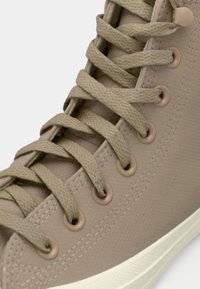 Beiger High-Top-Sneaker mit strukturiertem Gummiobermaterial, flachen Baumwollsenkeln und runden Ösen; verfügt über eine weiße Gummisohle.