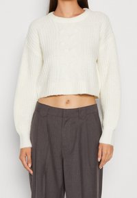 Weißer gestrickter, verkürzter Pullover mit gerippter Struktur, runder Ausschnitt und Zopfdetails in der Mitte, kombiniert mit grauen, hochtaillierten Hosen.