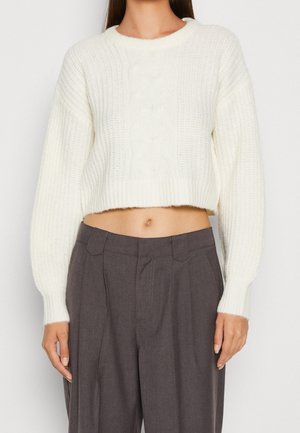 Witte gebreide cropped trui met ribbelstructuur, ronde halslijn en kabeldetails in het midden, gecombineerd met grijze high-waisted broek.