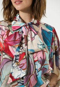 Blusa de seda con un diseño floral en colores vibrantes, con un cuello de lazo atado y mangas fluidas, que muestra una textura suave y patrones intrincados.