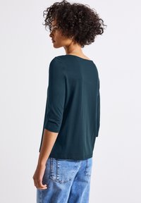 Mörk teal tröja med trekvartsärmar och bred halsringning, i kombination med ljusa denimjeans. Slätt tyg med avslappnad passform och enkelt design.