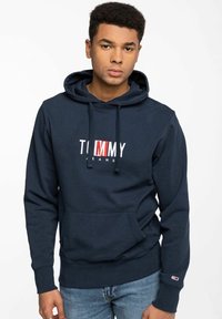 Sudadera de color azul marino hecha de algodón, con un bolsillo frontal, capucha con cordón y el logo "TOMMY JEANS" bordado en blanco y rojo en el pecho.