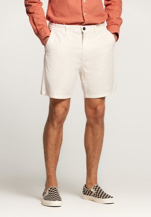 Lichtbeige katoenen shorts met riemlussen, voorzakken en een enkele knoopsluiting, gecombineerd met geruite zwart-witte instapschoenen.