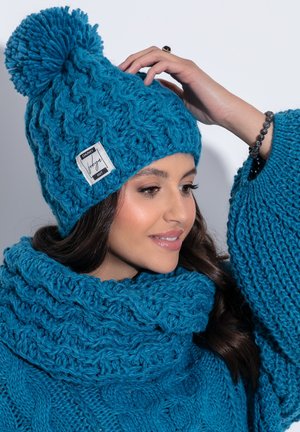 Gruby, turkusowy dzianinowy kapelusz z dużym pomponem i naszywką. W połączeniu z teksturowanym, turkusowym szalem w nieskończoność, obydwa z misternymi wzorami.