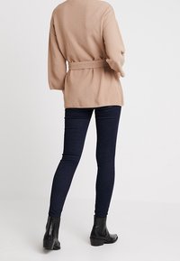 Beige stickad omslagströja med långa ärmar och bindebälte, kombinerad med mörkblå skinny jeans och svarta ankelstövlar.