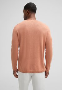 Maglione a maniche lunghe in morbido tessuto a trama pesca. Presenta un colletto tondo, polsini a coste e una vestibilità rilassata, indossato con pantaloni chiari.