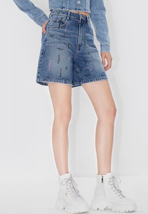Shorts en denim à taille haute avec un ourlet cousu, présentant des motifs de texte imprimés en noir et rose sur un tissu bleu clair. Associés à des bottines blanches.