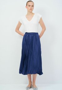 Jupe midi plissée marine avec une taille élastique, associée à un T-shirt blanc à col en V et des ballerines gris clair. Tissu lisse et léger.