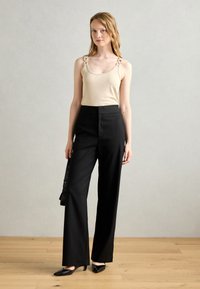Beige tank top with decorative chain straps and black high-waisted wide-leg trousers. Matte texture and sleek silhouette. Black heels.