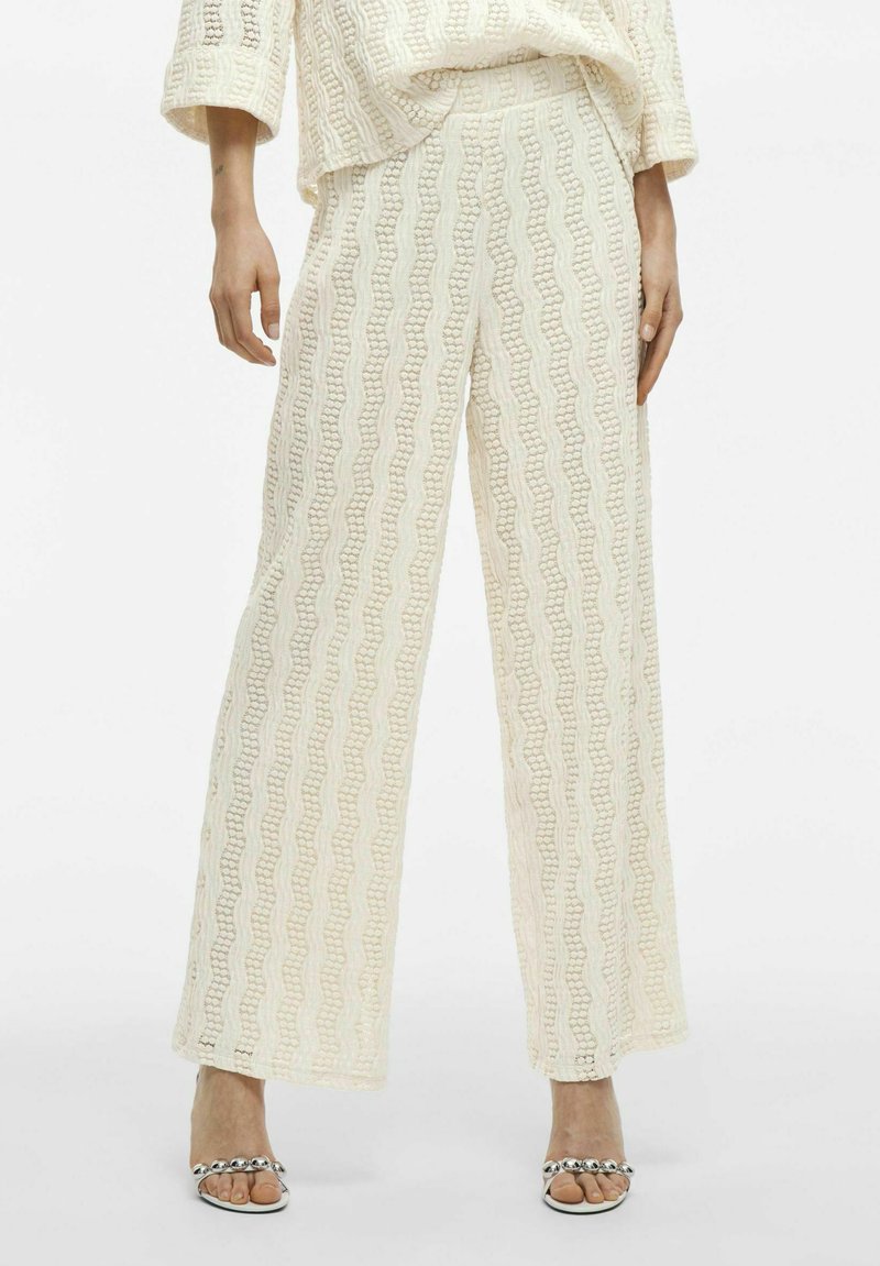 Pantalones de pierna ancha en tejido crema texturizado con patrones ondulados. La cinturilla es elástica y presenta un ajuste holgado para mayor comodidad.