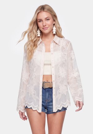 Donna bionda con grandi orecchini bianchi con nappe, camicetta trasparente con fantasia floreale e bottoni, crop top bianco, shorts di jeans blu e cintura beige.