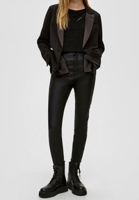 Blazer en satin noir sur un top noir pailleté, associé à un pantalon en simili cuir noir ajusté et des bottines de combat noires à lacets.