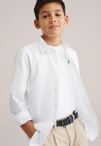 Cămașă albă cu nasturi peste un tricou alb, cu mânecile suflecate. Pantaloni deschiși la culoare și o curea neagră texturată completează ținuta.