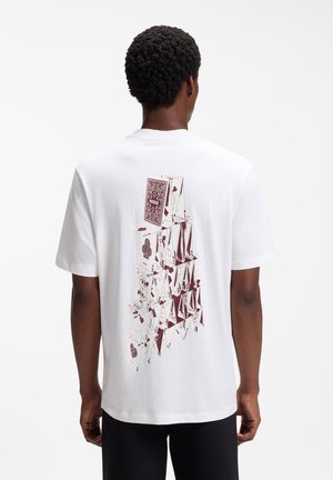 Mand iført en hvid t-shirt med et detaljeret mørkerødt hus af spillekort på ryggen, stående mod en ensfarvet hvid baggrund.