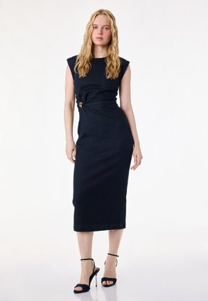 Kvinde med langt blondt hår iført en ærmeløs, figursyet navy midi-kjole med en side spænde og sorte højhælede sandaler, stående mod en hvid baggrund.