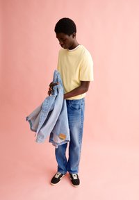 Gelbes T-Shirt, blaue Jeansjacke und blaue Jeans, gehalten von einer Person. Schwarze Sneakers mit weißen Schnürsenkeln. Sanfter rosa Hintergrund.