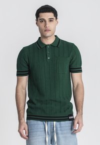Polo de punto verde con un patrón texturizado, de mangas cortas y un bolsillo en el pecho. Presenta acentos de rayas en el cuello y los puños.