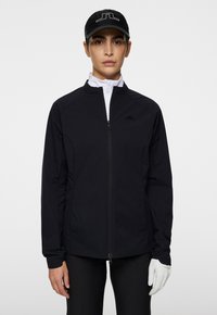 Svart idrottsjacka med dragkedja fram, uppstående krage och elastiska muddar. Vit inre lager synligt vid halsen. Vit handske på handen.