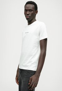 Camisa T-shirt branca de mangas curtas feita de algodão, apresentando um decote redondo e um pequeno logótipo preto "Replay" no peito. Design simples, estilo minimalista.