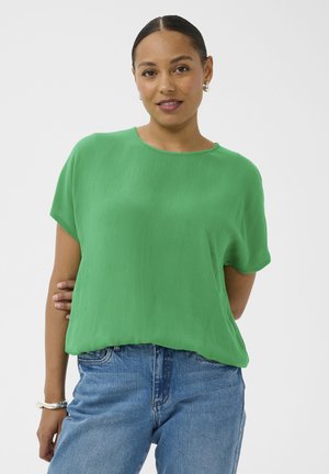 Jeune femme aux cheveux tirés en arrière portant un chemisier vert à manches courtes et un jean bleu, posant avec une main dans le dos.