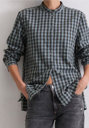 Person wearing a blue and black checkered button-up shirt with sleeves rolled up and hands in pockets of faded black jeans.