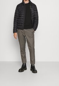 Veste matelassée noire, légère et matelassée, associée à un pull foncé, un pantalon à motifs et des bottines noires. Debout devant un fond uni.