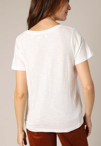T-shirt blanc à manches courtes en tissu léger, avec un col rond et un petit détail de bouton au dos.
