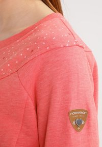 Felpa color corallo con pannello sulle spalle in tessuto testurizzato, caratterizzato da pois bianchi, e una patch con logo in pelle marrone sul manica.