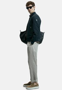 Dunkelmarineblaue Bomberjacke mit einer Reißverschlusstasche, kombiniert mit hellbeigen Hosen und braunen Slippern. Das Modell trägt eine schwarze Lederhandtasche.